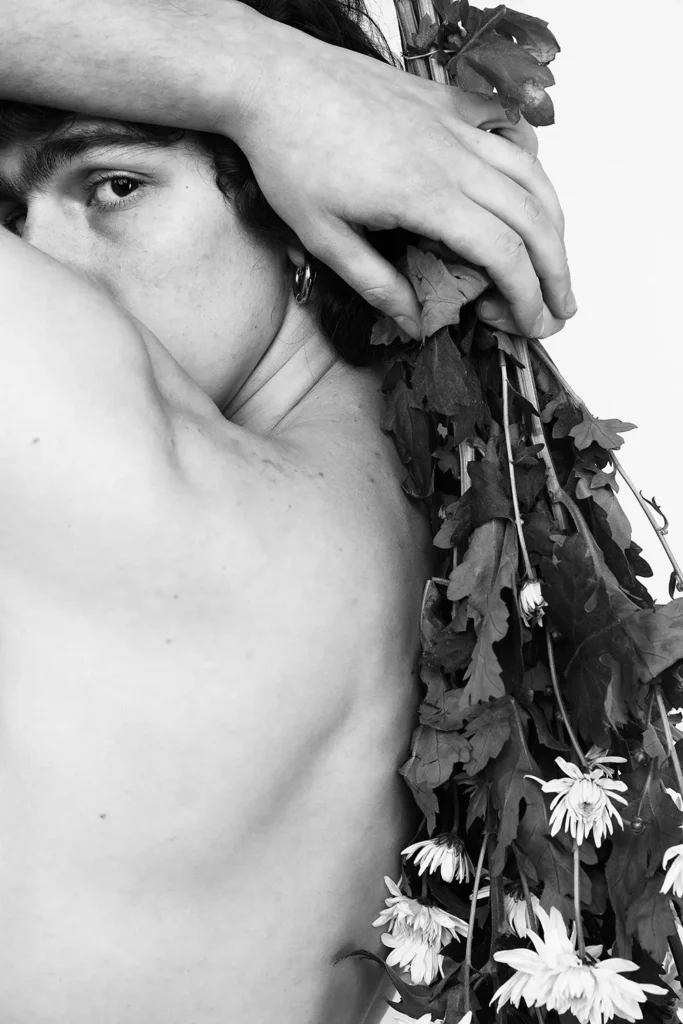 Retrato en blanco y negro de un hombre con el torso desnudo que cubre su rostro con un brazo. Margaritas secas y hojas cuelgan de su espalda.