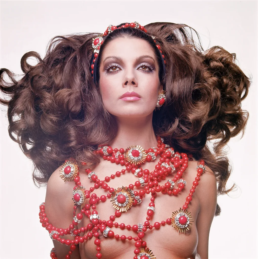 Ritratto in studio di una modella con gioielli Valentino rossi e dorati e capelli voluminosi, fotografata su sfondo bianco.