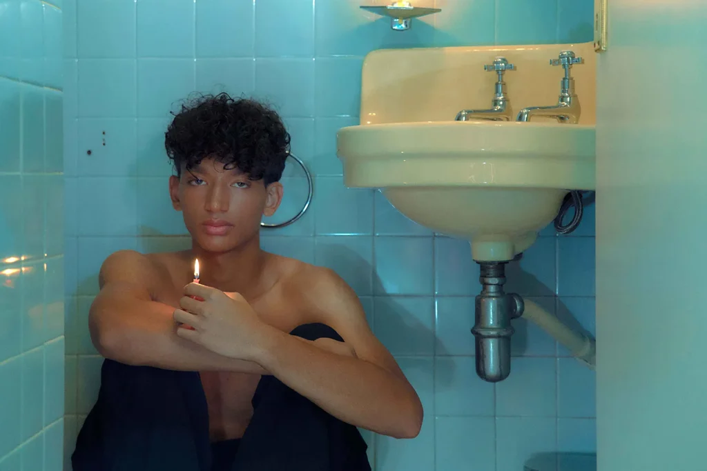 Young man sitting shirtless in a tiled bathroom holding a lit lighter under a sink