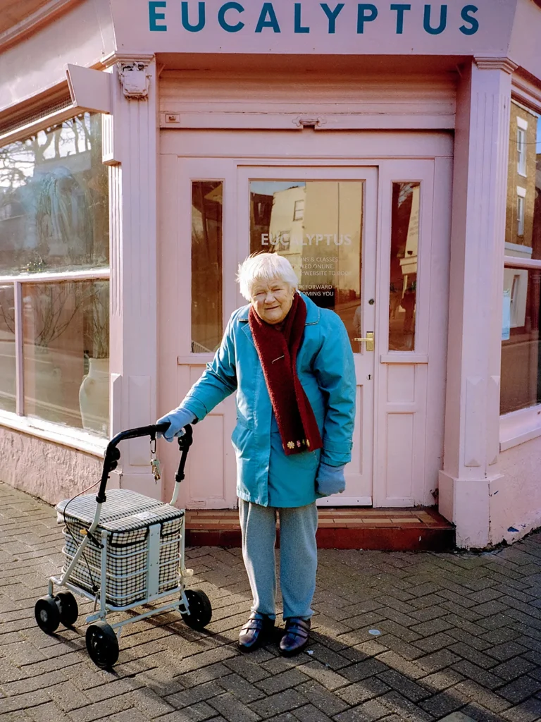 Elderly woman in blue coat standing with walker in front of pink storefront labeled "Eucalyptus"