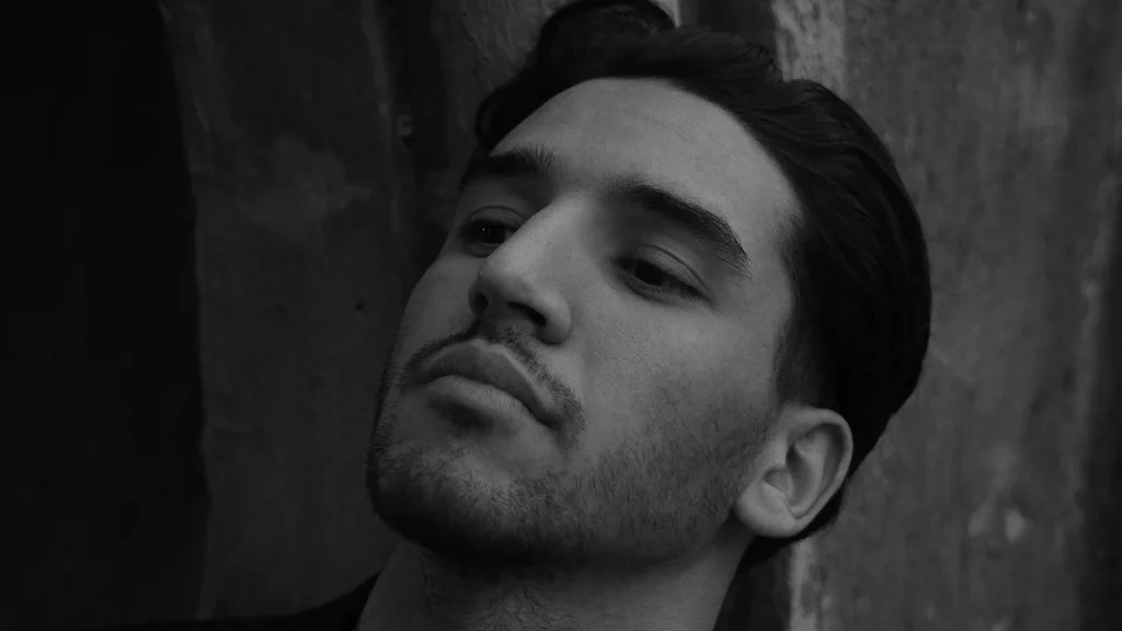 Close-up portrait of a man leaning against a wall in soft shadow, eyes half-closed.
