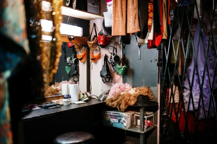 Backstage room with hanging costumes, wigs, and glittering accessories — the faded glow of a show that has ended.