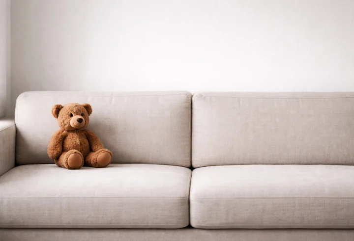 Small brown teddy bear sitting on the left side of a neutral sofa against a white wall